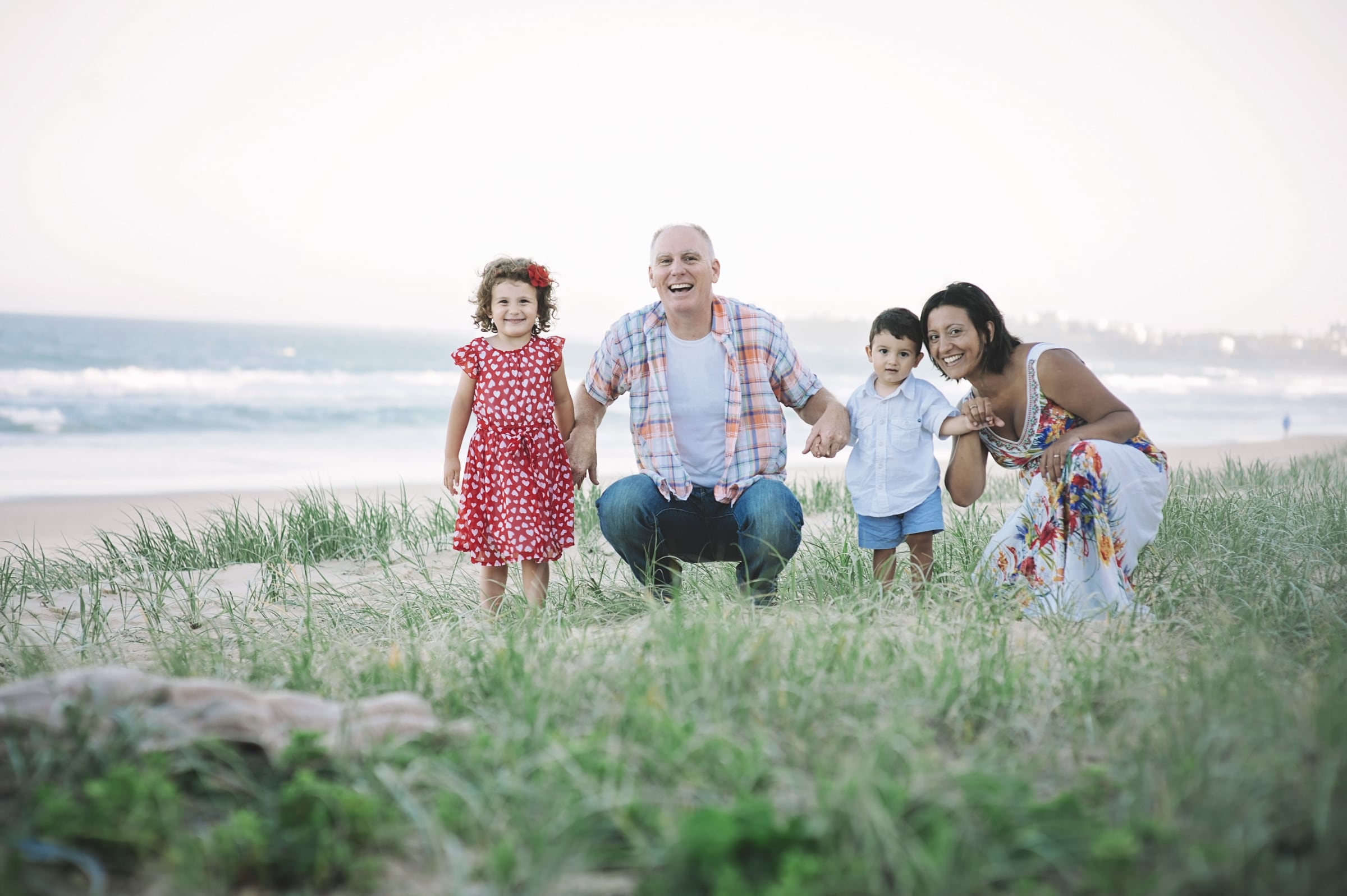 Rob and Silvina crouched in the dune grass with their two young children, all smiling, the ocean and a hazy sky behind them at dusk.